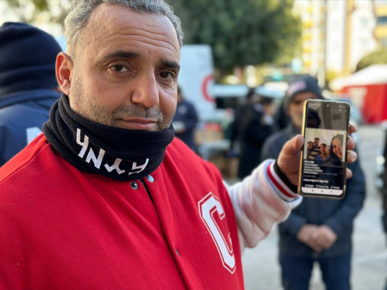 A man in a red jacket holds up a mobile phone with a photo of fur people on it.
