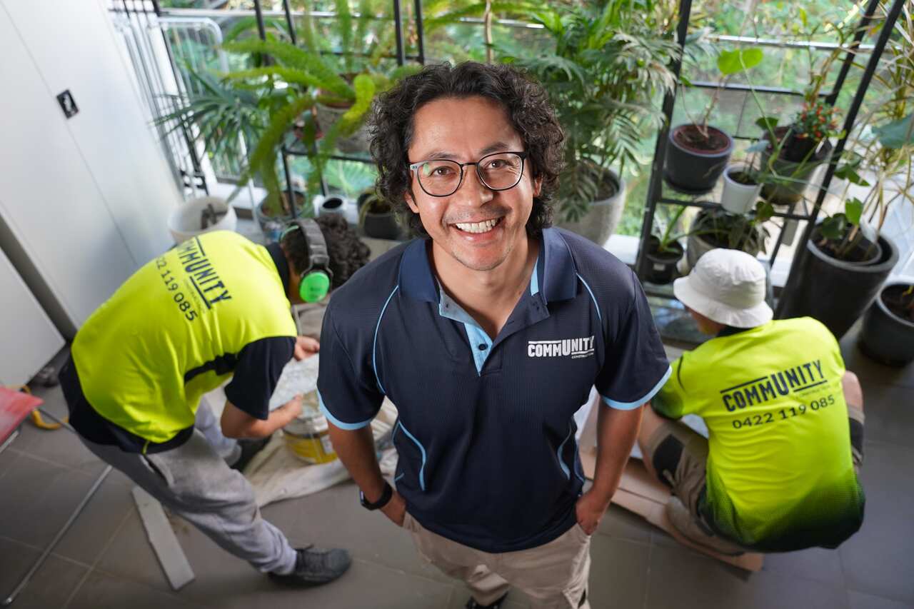 A man in glasses and a navy polo smiles into the camera, while two construction workers kneel behind him.