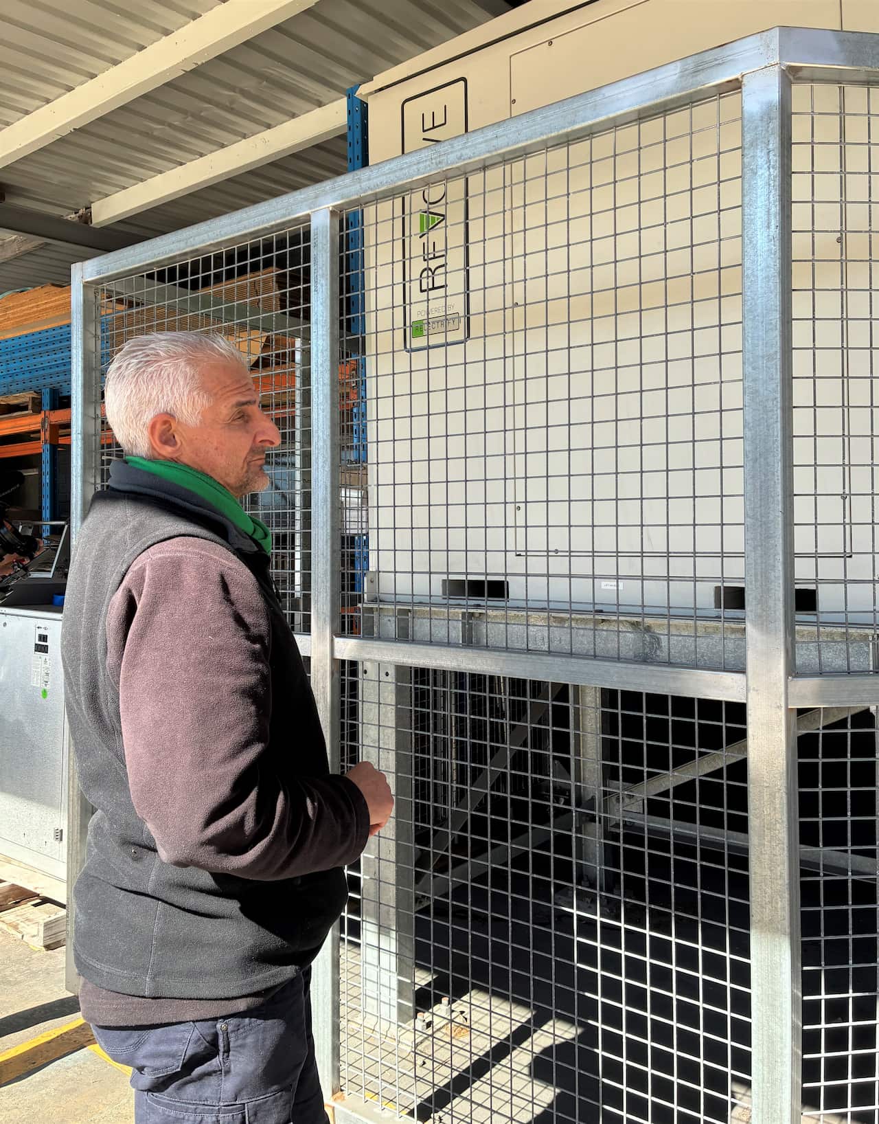 A man in a grey sweater stands next to a wire cage, looking at a battery housed inside.