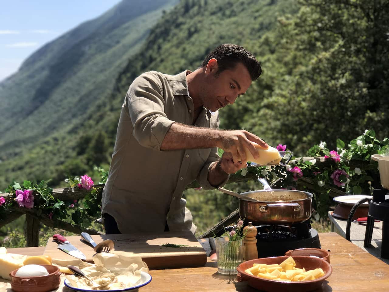 A man stands outdoors at a table, with mountain views behind him. He grates cheese into a pot on a portable cooktop on the table.