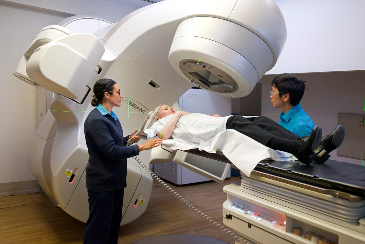 A woman is being treated by a radiation therapy machine. There are two medical personnel standing next to the woman on the machine on either side of her. 