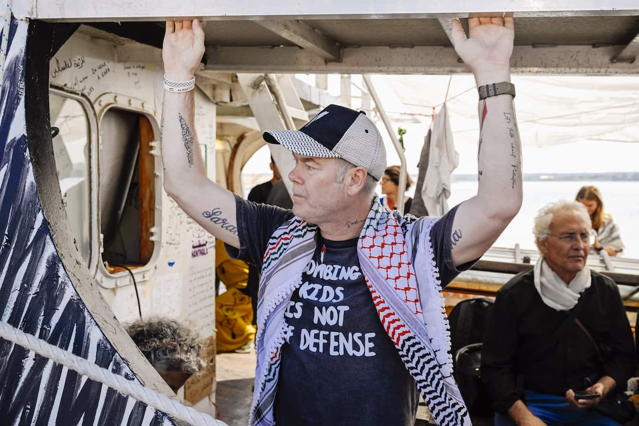 A man holds onto the side of a ship. He is wearing a blue t-shirt with white writing on it that reads: Bombing kids is not self defense