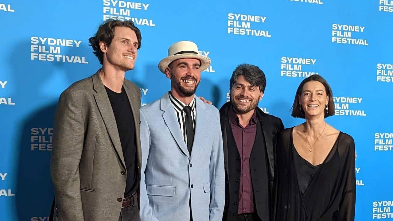 Three men in suits and a woman in a black dress stand in front of a blue wall that has "Sydney Film Festival" written all over it in white.