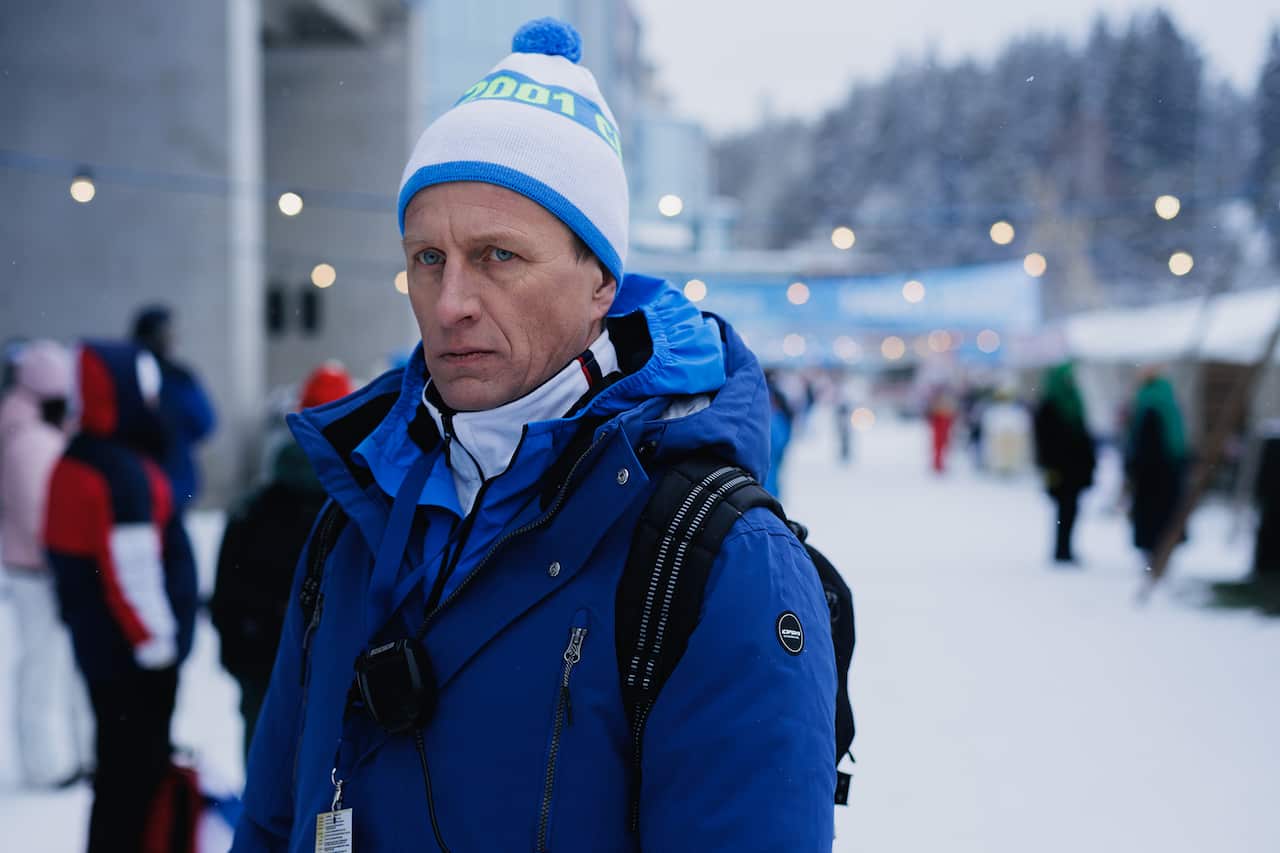 A man in a blue jacket and white beanie, with an identity pass handing around his neck, stands in a snowy street.
