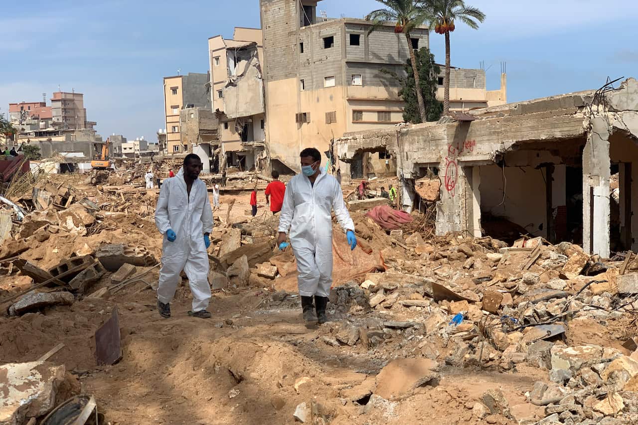 Two men in white hazmat suits and blue gloves walk through the mud and ruin of Derna. The man on the right wears a face mask.