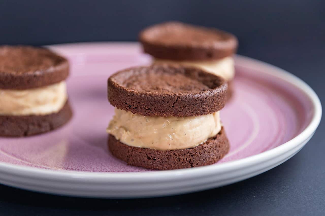 An ice-cream sandwich, with caramel ice-cream between two chocolate biscuits, sits near the edge of a wide pink plate. Two more stacks can be seen behind, sitting on the same plate. 