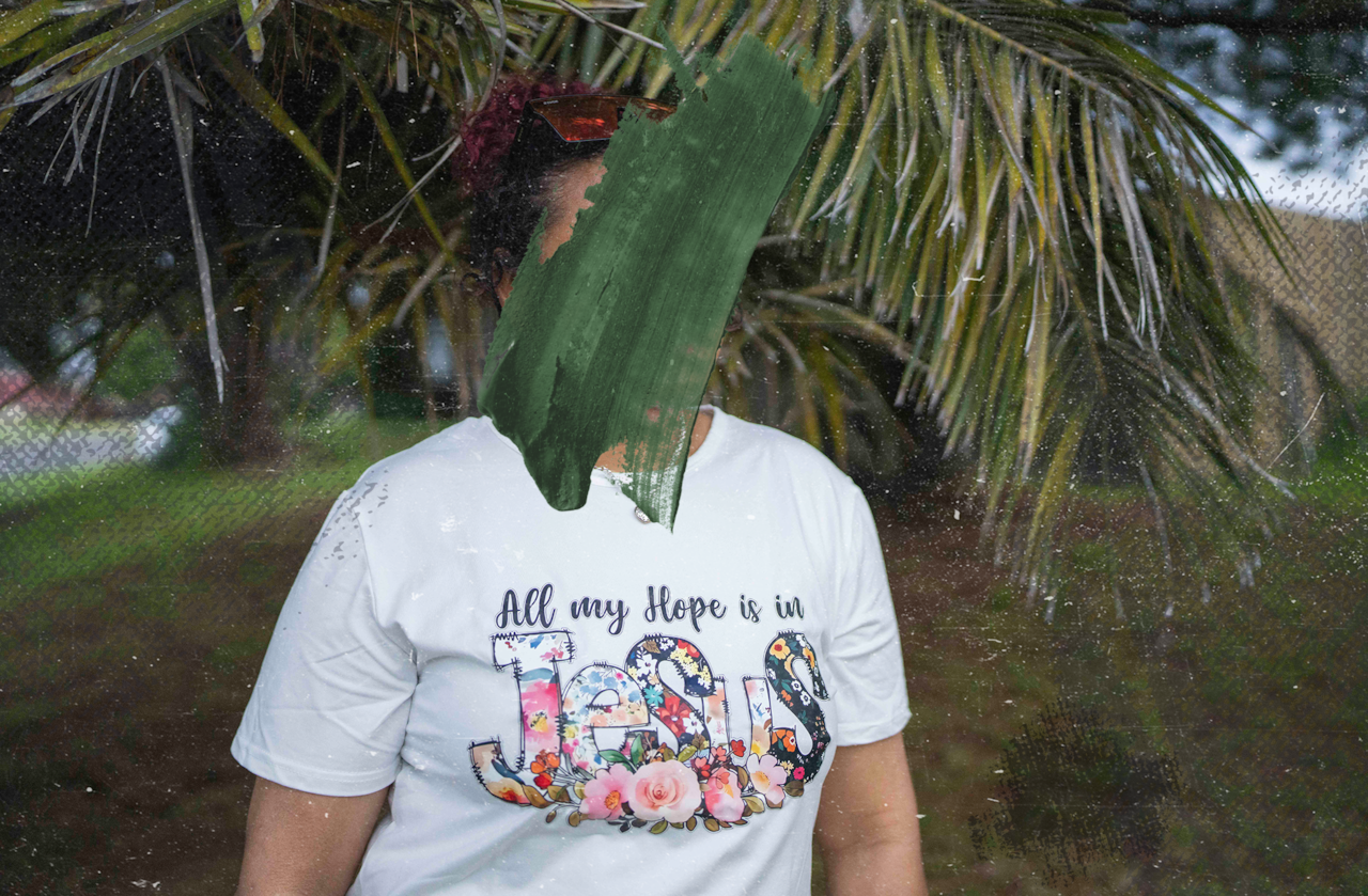 A woman is standing outside near a palm tree and wearing a white t-shirt that reads: "All my hope is in Jesus". Her face is obscured by a black smudge.