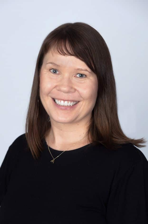 A person with medium-length dark hair smiles in front of a blank wall