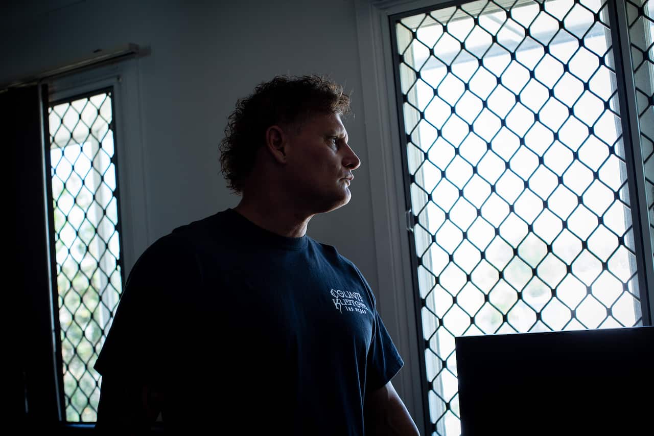 A middle-aged man with short curly hair wears a black t-shirt and looks out of a barred window.