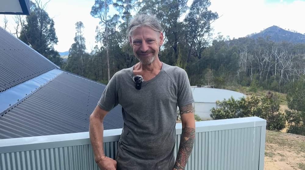 A man smiling, standing on a balcony of a house surrounded by trees.
