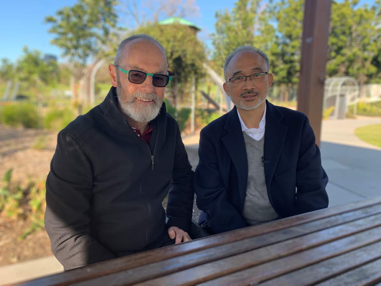 Two men in their 60s, one Australian and one Vietnamese, sitting on a park bench.