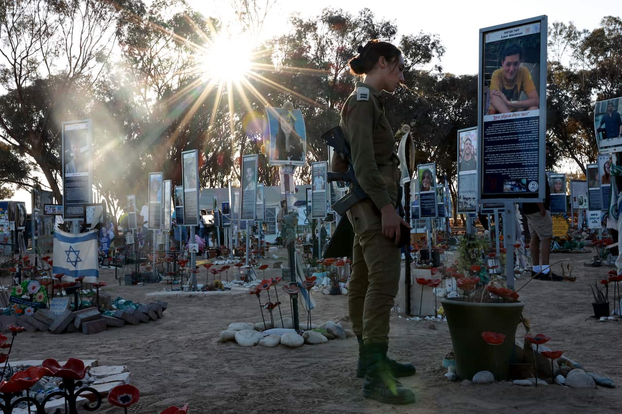 A woman in military garb stands in front of a sign showing a victim of the October 7 attack in Israel. Many other placards are visible around her, each with flowers, images and writing about the victims.