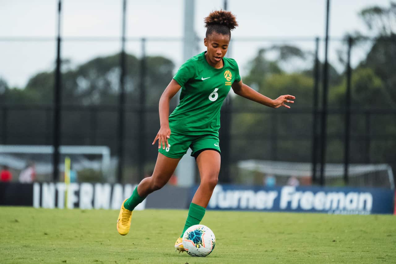 A young woman in a green football kit kicks a ball on the pitch.