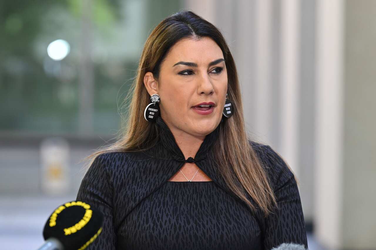 Lidia Thorpe speaks outside. There is a microphone to her right. She is wearing all black, and has earrings which read 'sovereignty never ceded'. 