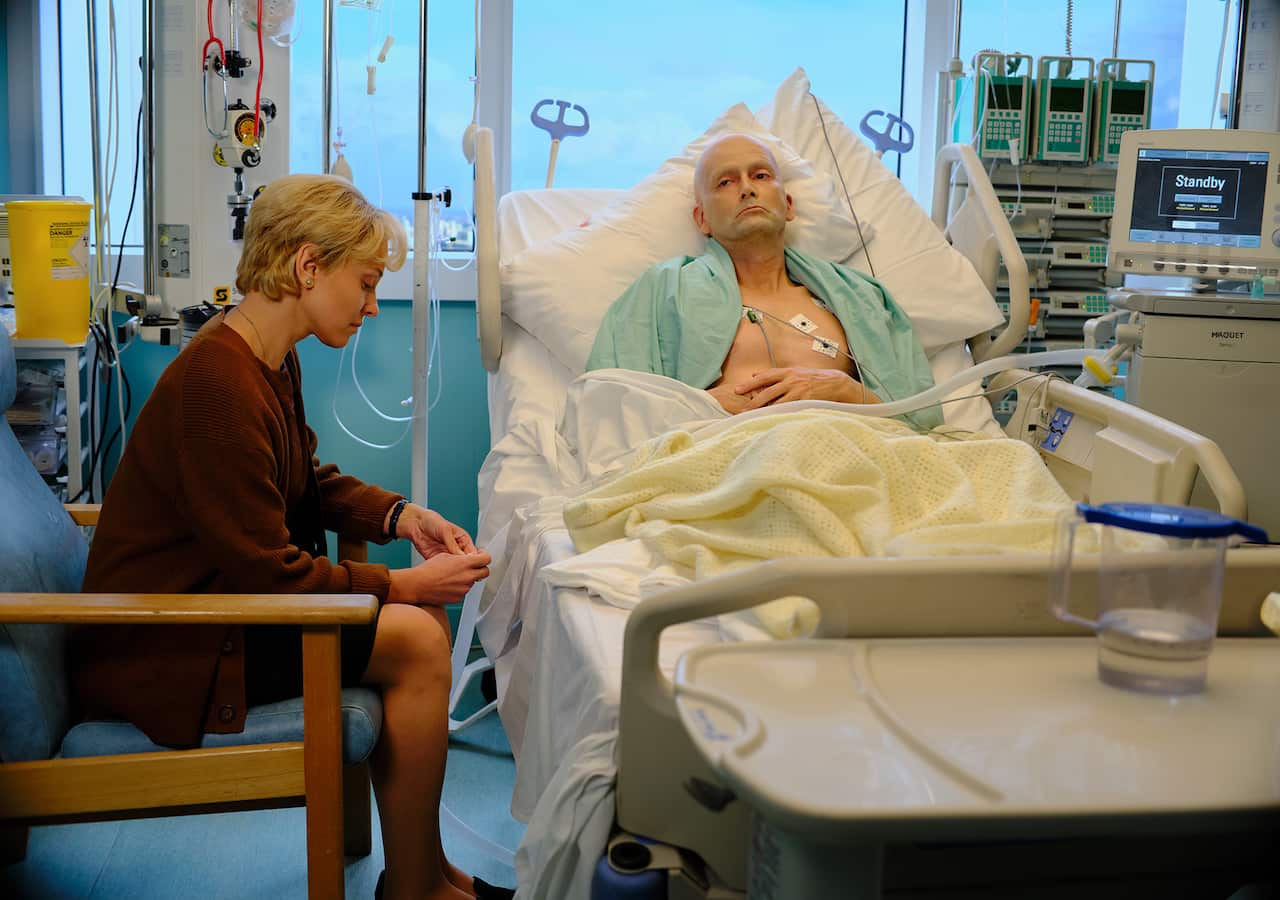 A pale, ill-looking man lies in a hospital bed. A woman sits in a chair by the bed, looking down at her hands. 