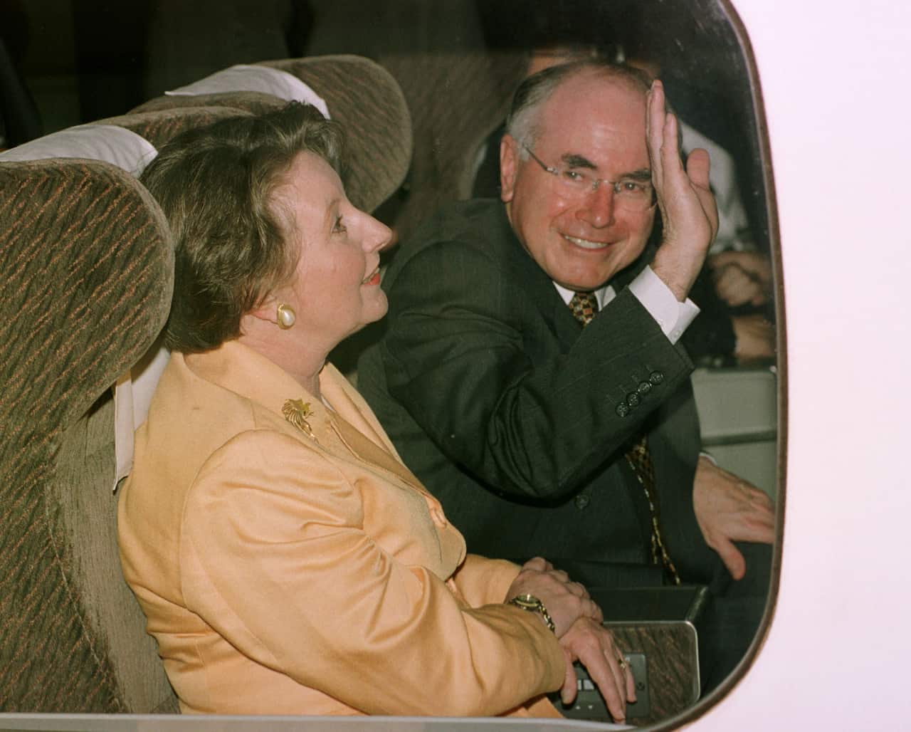 A man in a black suit waves as he sits next to a woman in a yellow full sleeves top on a train.