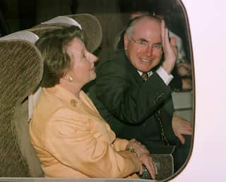A man in a black suit waves as he sits next to a woman in a yellow full sleeves top on a train.