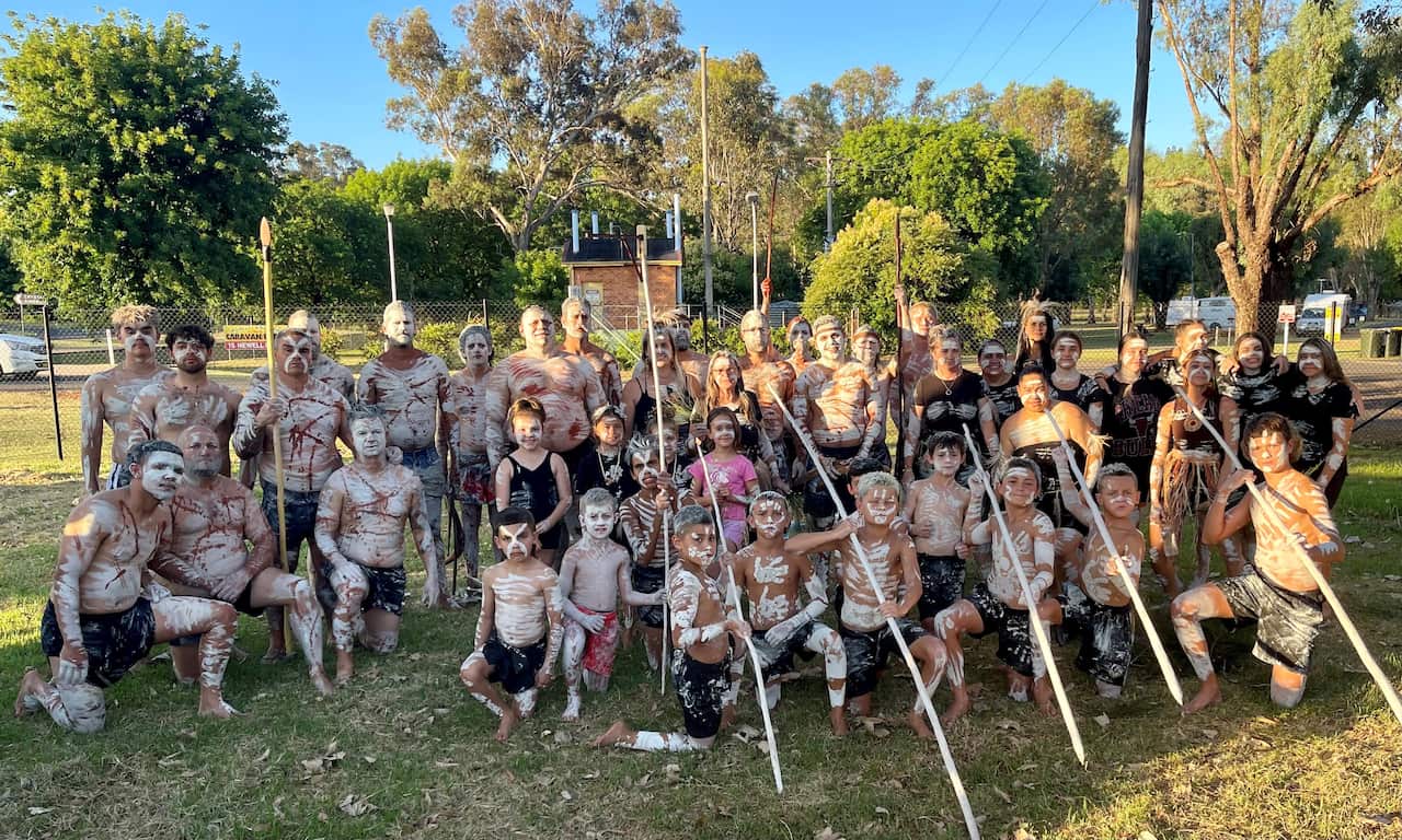 A group of around 50 Gomeroi men, women and children standing and kneeling, all painted in cultural white ochre, and some holding white spears, as they prepare to dance in support of the local community's fight against a major gas development.