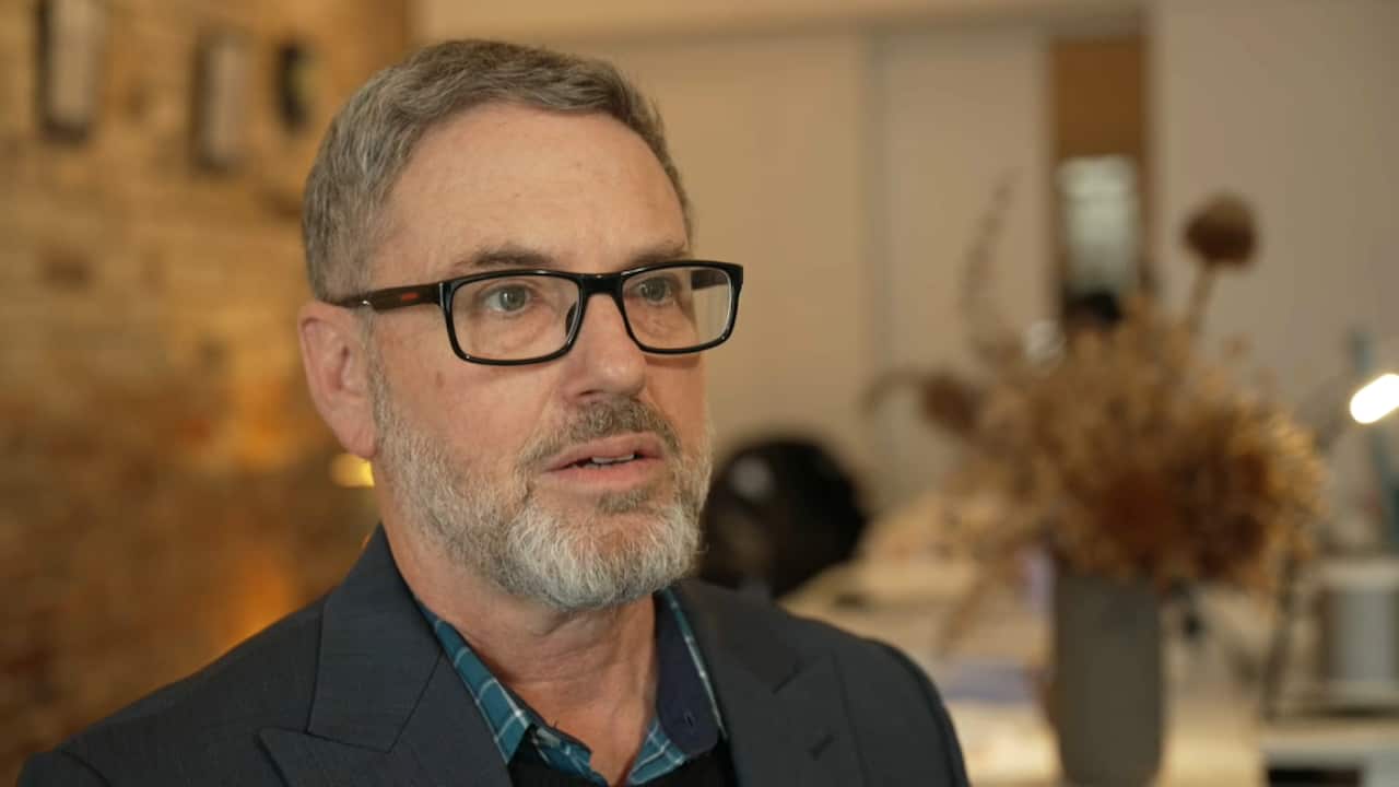A man with a white goatee beard and glasses stands in a suit jacket
