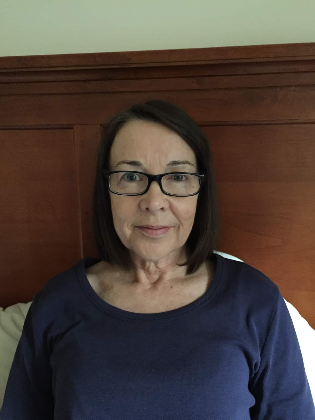 A woman in glasses sits in bed with a neutral expression on her face.