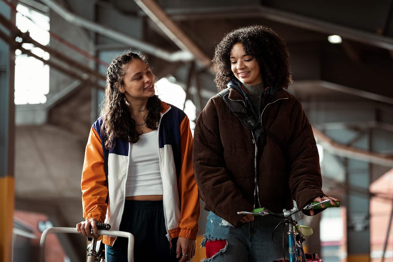 Two women stand beside each other, smiling, in what looks like a semi-industrial street. They are both holding the handlebars of bikes. 