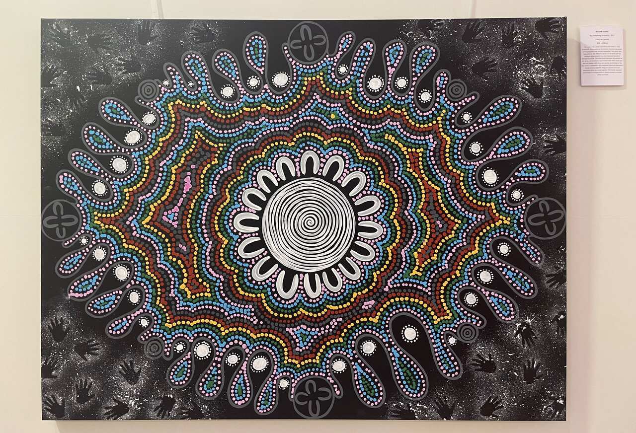 An Aboriginal dot painting with black background and multicoloured dots in an intricate design is hung up on a canvas on the wall. 