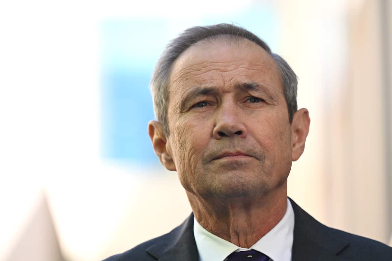 Roger COok, in a black suit, white shirt and dark tie, standing in front of a white building.