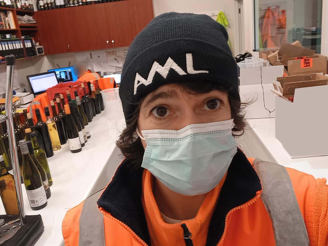 A woman wearing a face mask takes a photo in front of a room filled with wine bottles