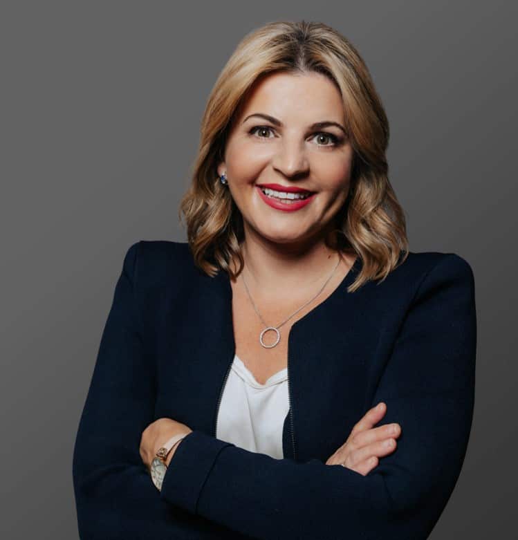 A woman wearing a navy blue suit jacket and a white t-shirt smiles at the camera.