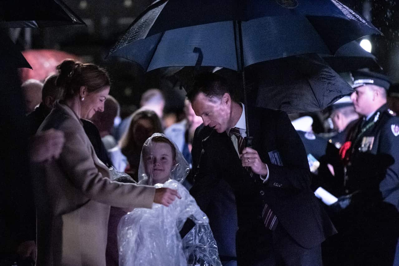 A man in a black suit and tie holding an umbrella stands near a young child wearing a rain poncho and a crowd of people.