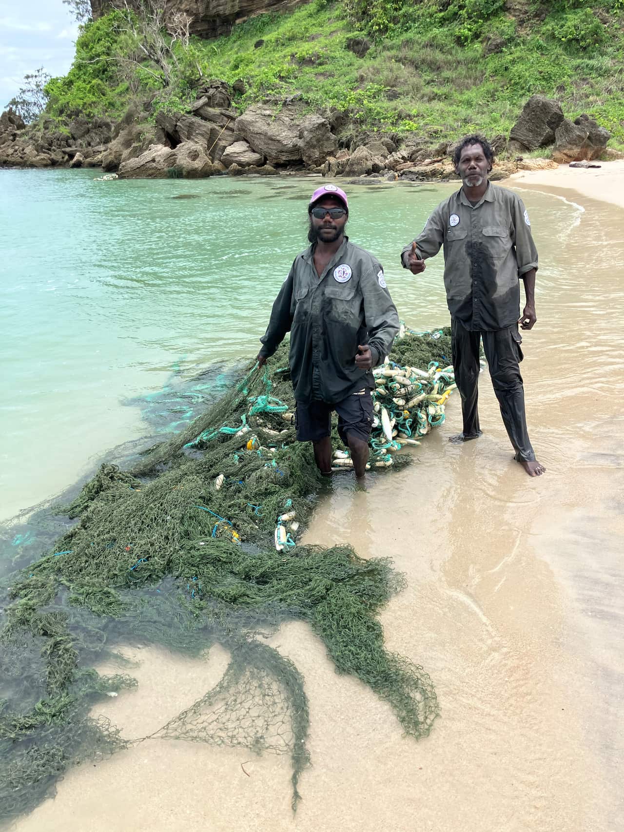 two male Indigenous rangers pulling a large fishing net with rubbish attached in to shore.