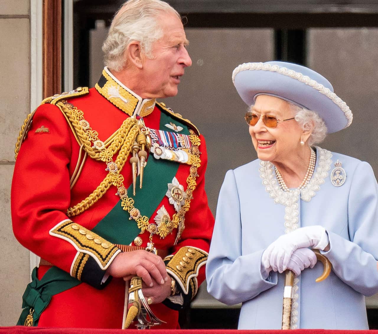 Prince Charles wearing red military uniform standing next to Queen Elizabeth II who is wearing a light blue coat 