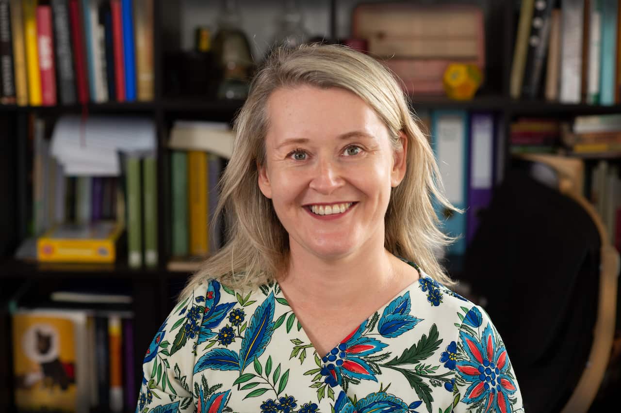 A woman with shoulder-length blonde hair smiles in front of a bookshelf.