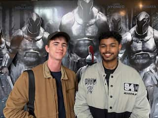 Two smiling young men pose side-by-side in front of a large movie poster featuring armoured, metallic figures.
