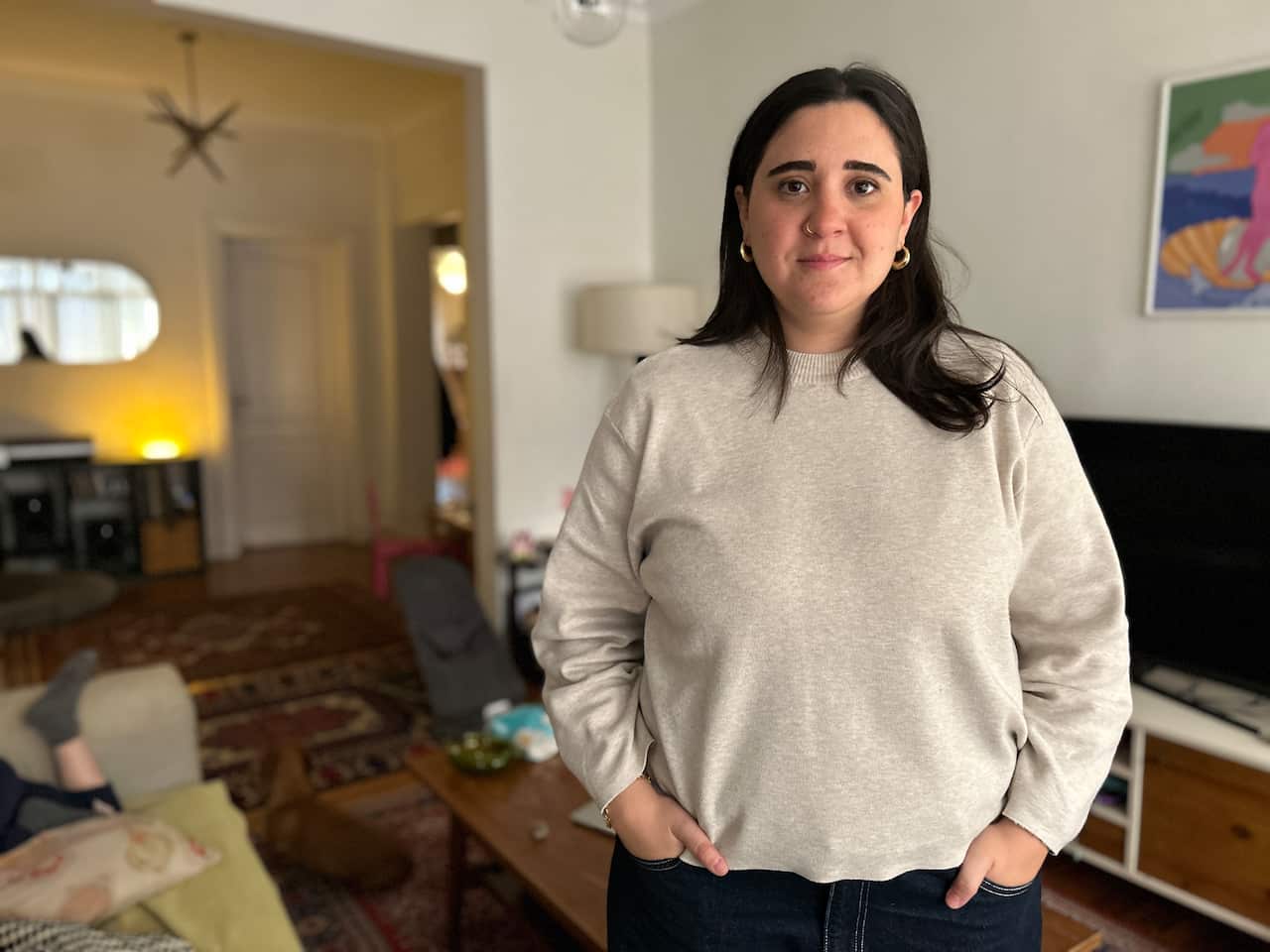 A woman with long dark hair stands in a cosy house wearing a grey jumper and with her hands in her pockets. She has a slight smile in her face.