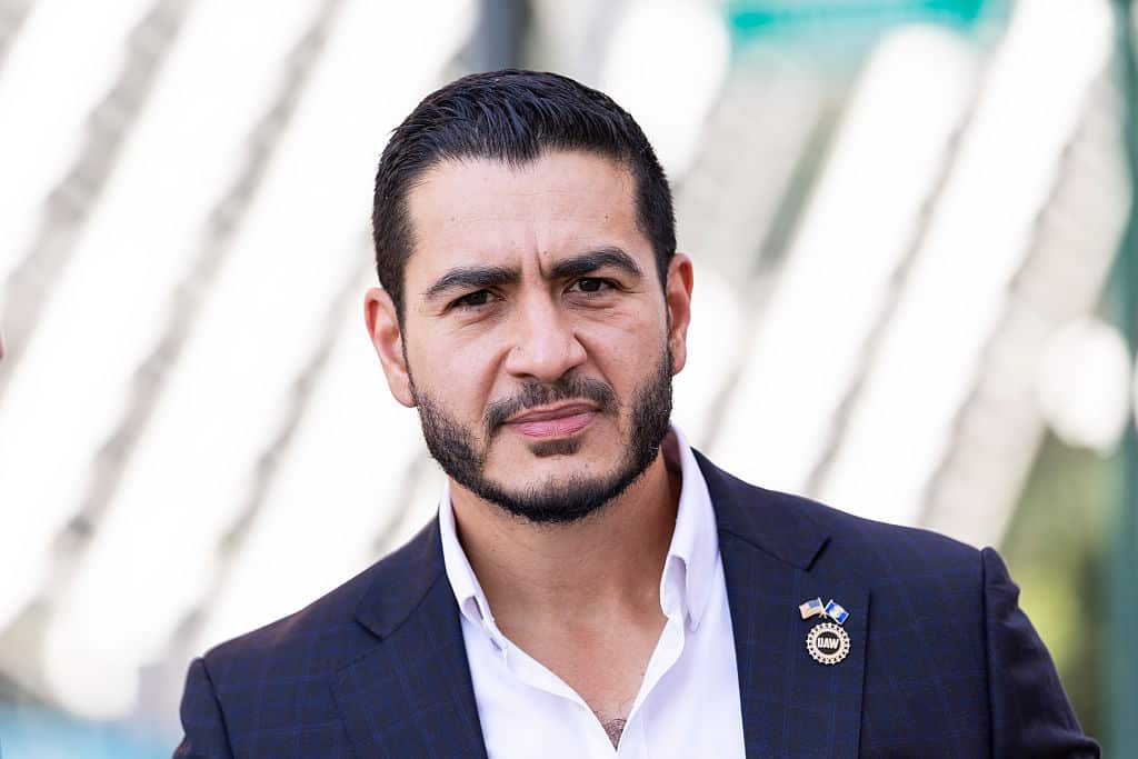 A man with dark hair and beard, and wearing a shirt and suit jacket, looks at the camera with a serious expression on his face.