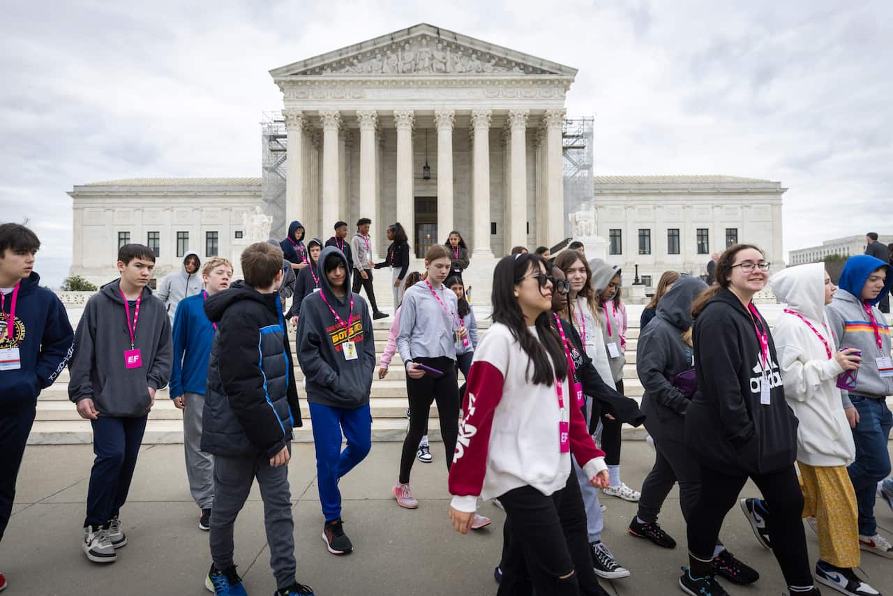 Group of students with lanyards around their necks walk past large, white US Supreme Court building.
