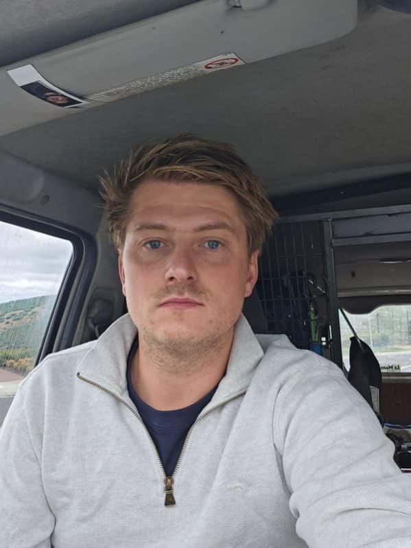 A white man in his thirties with blue eyes and a grey jumper on looks at the camera while sitting in his vehicle.