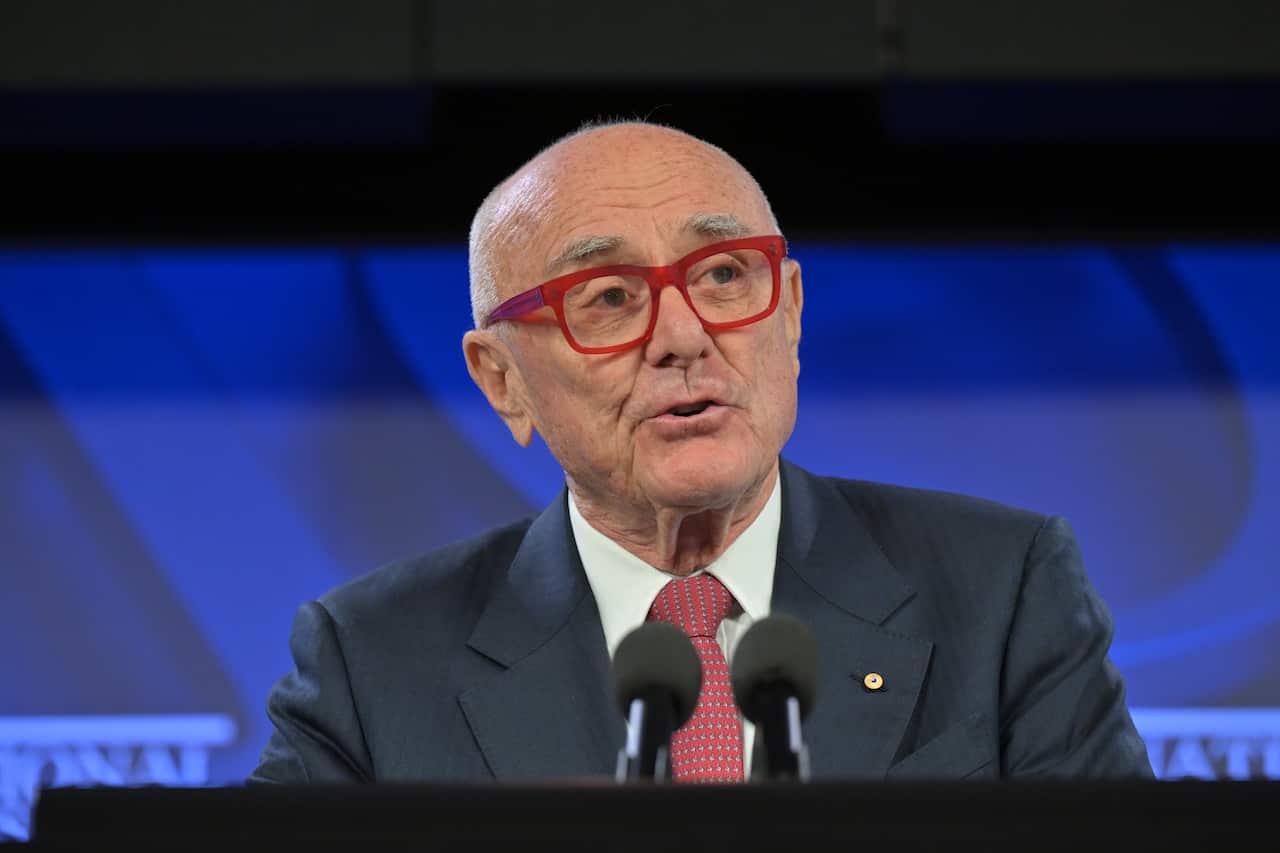 An older man with red glasses wearing a suit speaks into a lectern microphone 