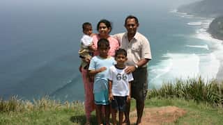 a mother and father with a baby and two young boys posing for a family photo on a headland with beach behind