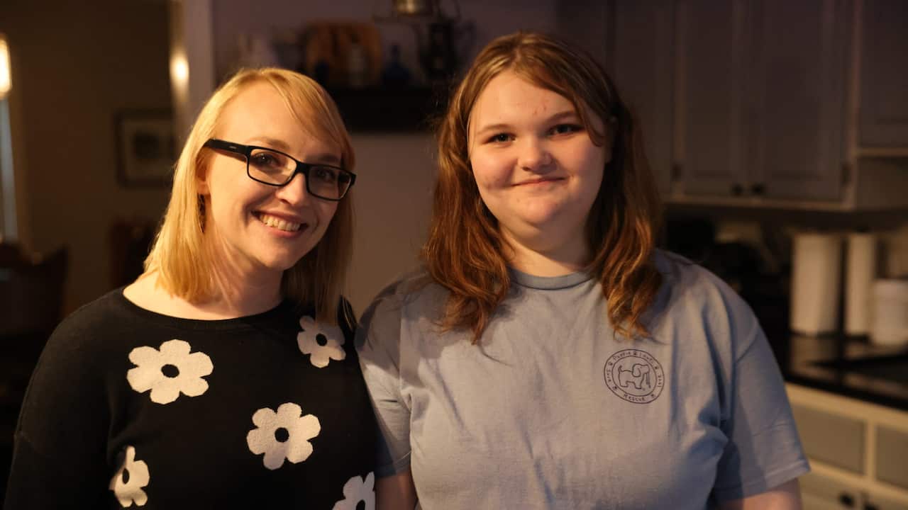 A blonde woman wearing dark-framed prescription glasses and a black sweater with a floral print stands next to a teenage girl wearing a grey t-shirt