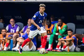 A footballer wearing a blue shirt and white shorts controls the ball on the pitch in front of teammates sitting on the sidelines bench.