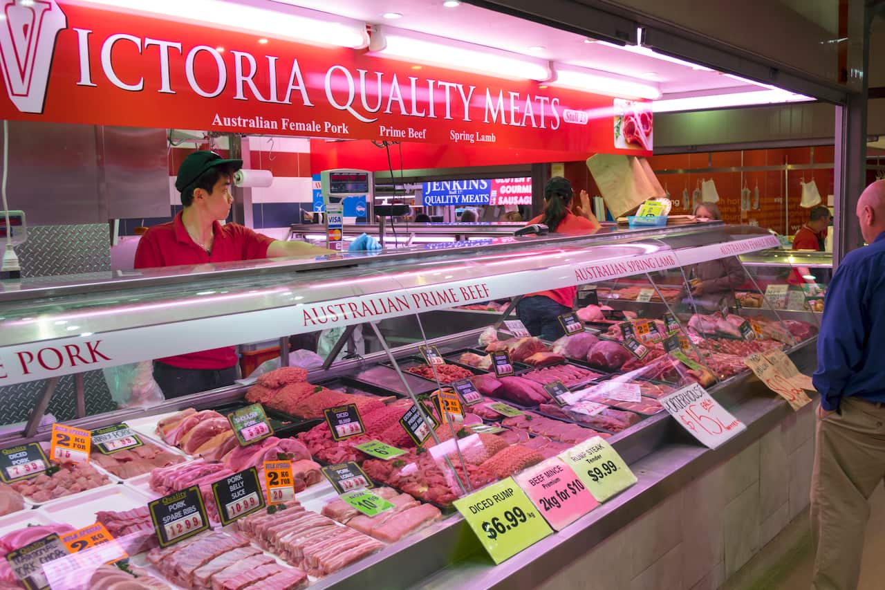 A salesperson wearing a red t-shirt stands behind a butcher meats display counter.