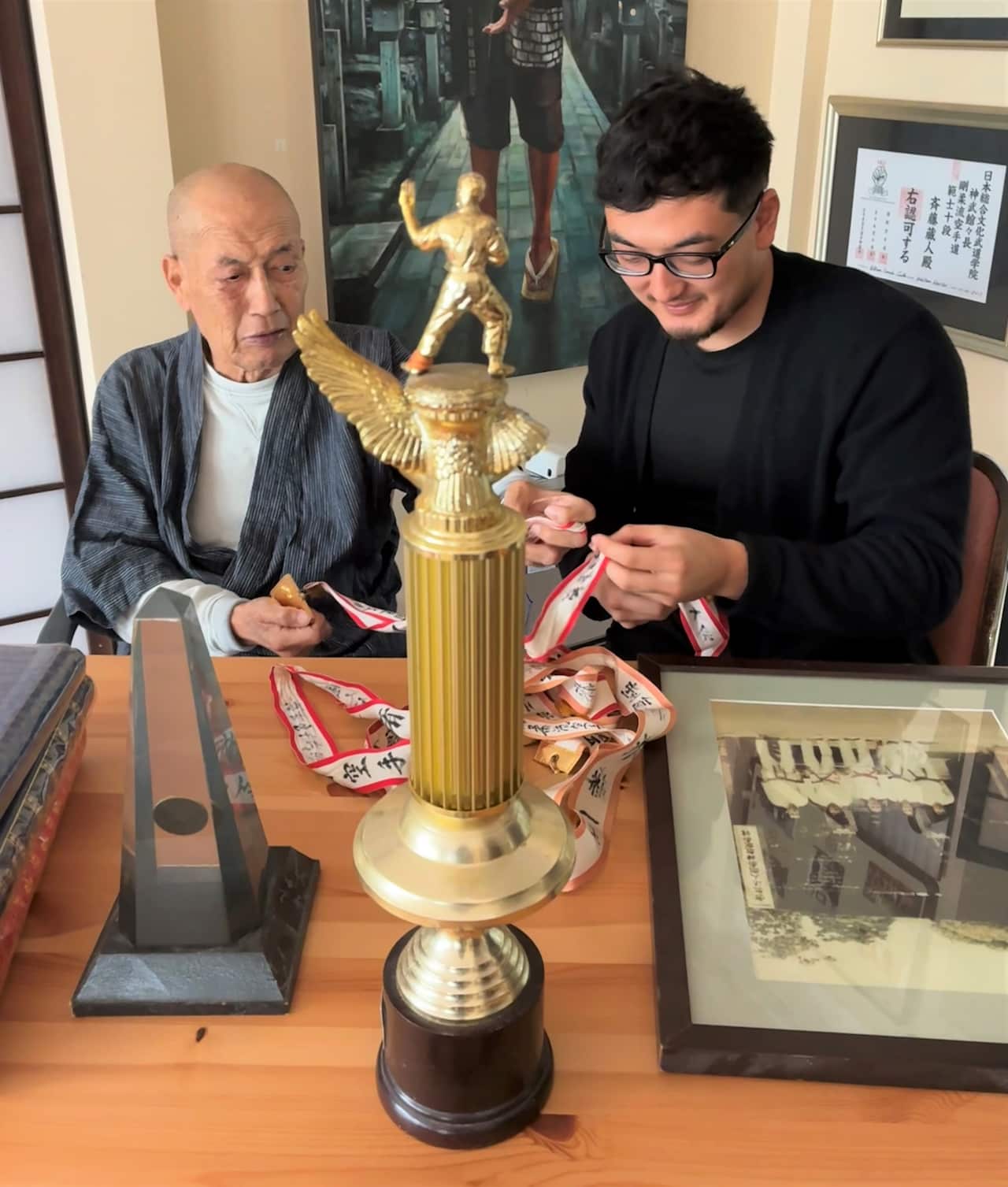 A man sitting at a table looks at medals and awards while an older man looks on.