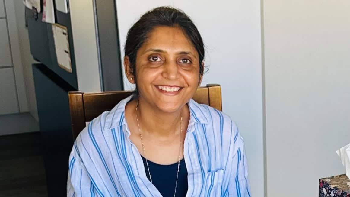 A middle-aged woman of South Asian descent wearing a white and blue striped shirt. She is smiling,