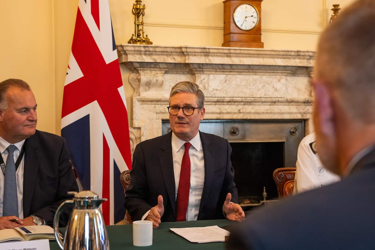 British Prime Minister Kier Starmer sits at the cabinet table with a group of others. Behind him is a Union Jack flag and a marble fireplace. 