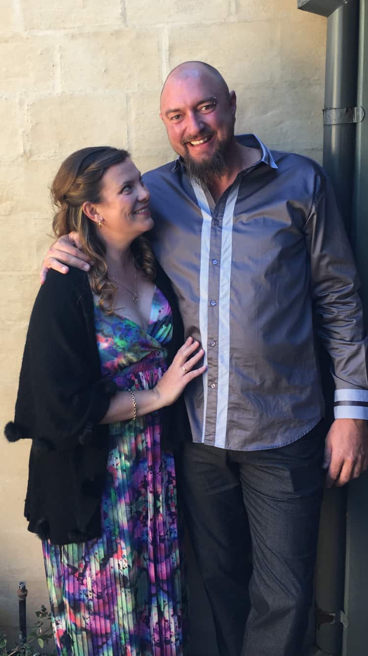 A couple stand and embrace happily, her looking at him and him looking at the camera.