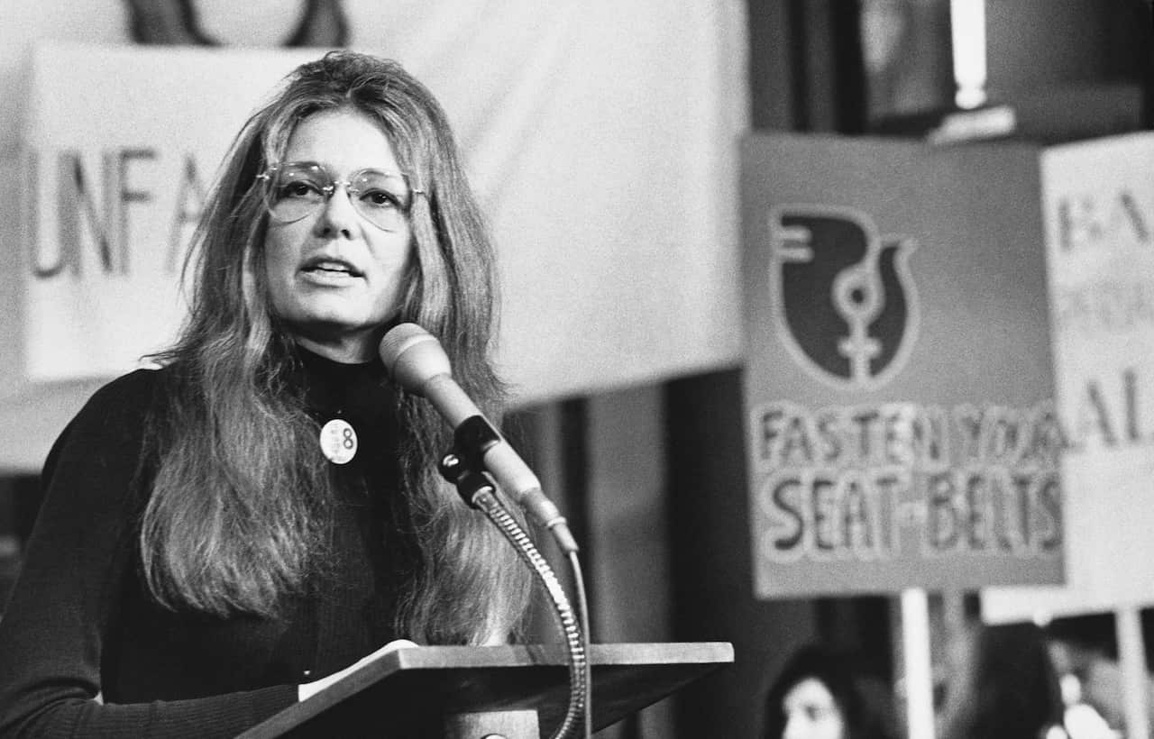 A black and white image of a young white woman speaking before a podium. People holding placards are behind her.