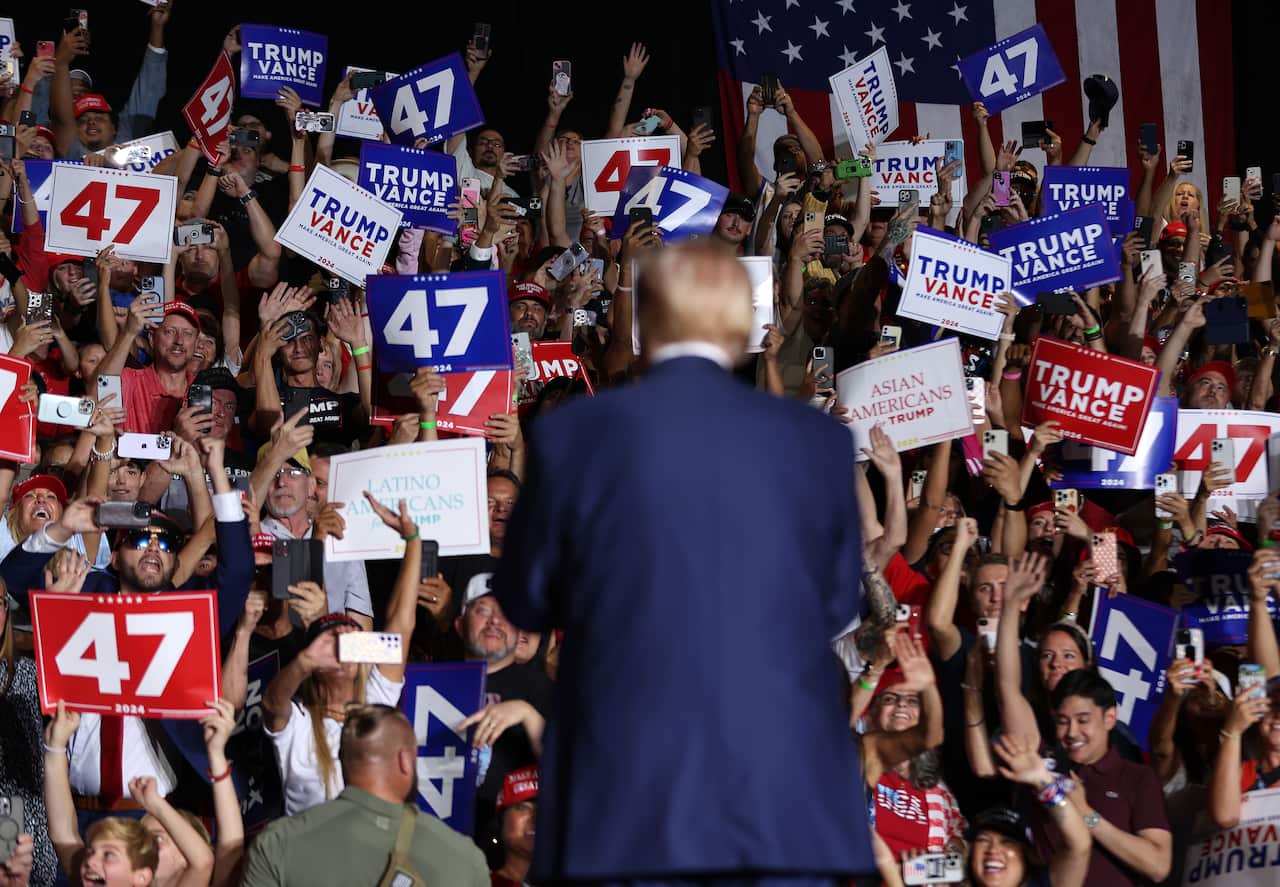 A man in a blue suit is addressing a rally. His back is towards the camera.