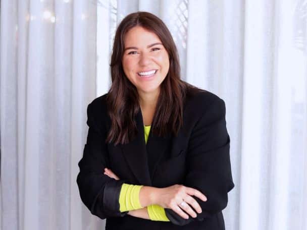 A woman with long brown hair is smiling while standing in front of a white curtain. She is wearing a black blazer over a neon yellow long-sleeved top.